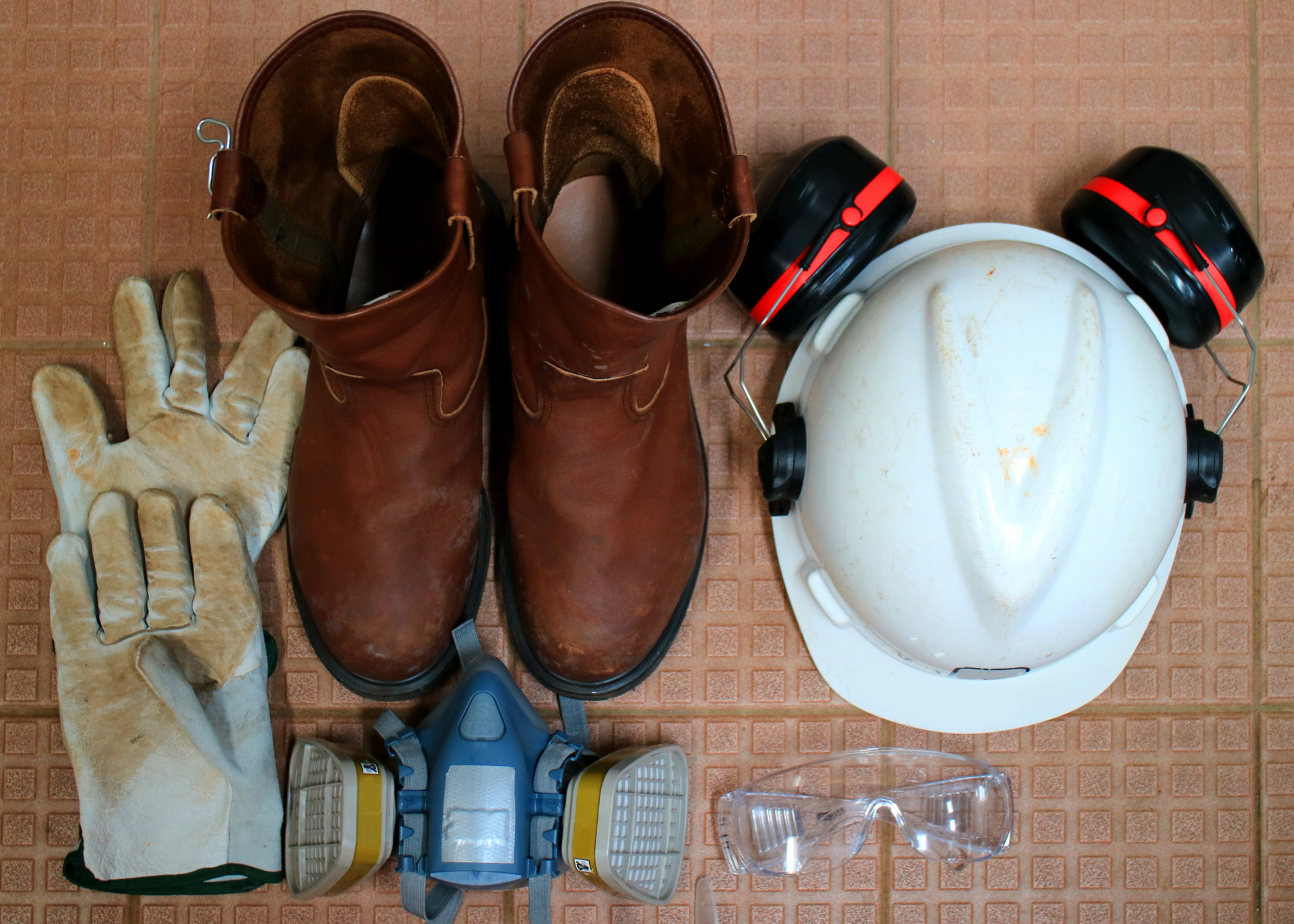 Brown work boots, gloves, respirator mask, hard hat, and safety glasses arranged on a mat