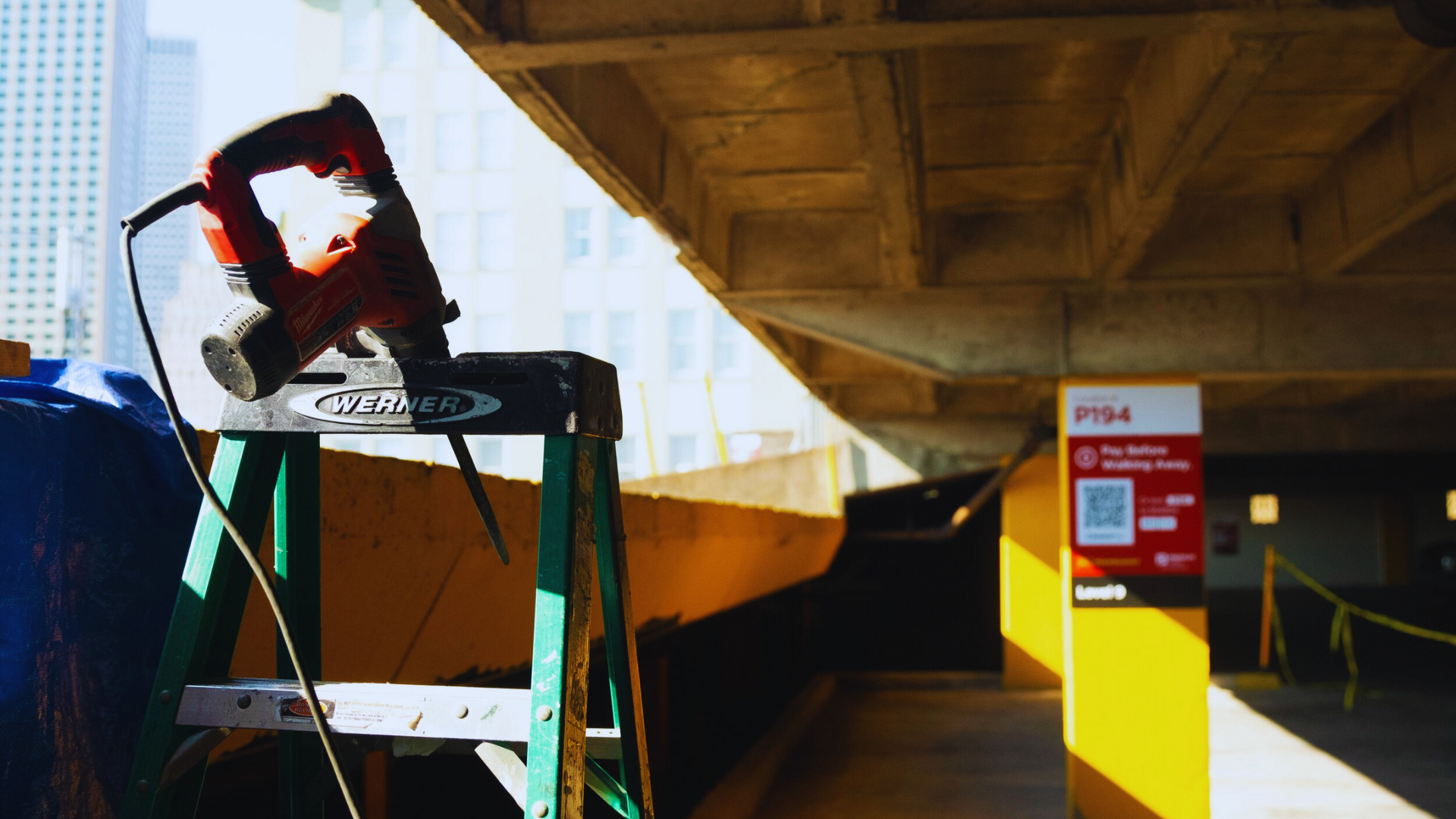 Parking garage entrance under a concrete overpass, with a broken sign on a metal frame in the foreground.