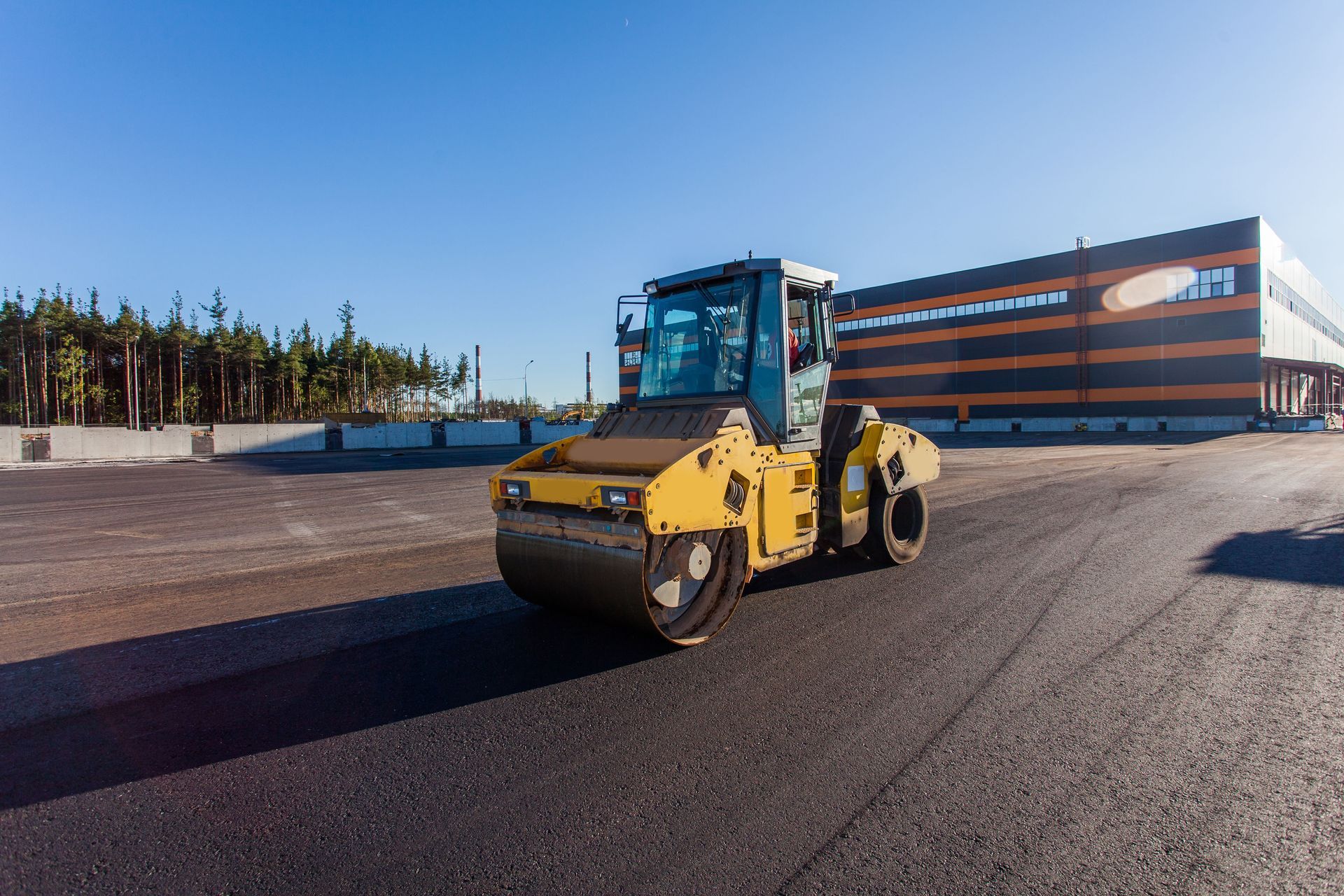 Yellow road roller on a paved construction site beside a modern building under a clear sky