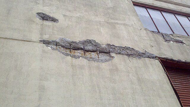 Cracked beige exterior wall with peeling plaster and a damaged horizontal section near the roofline