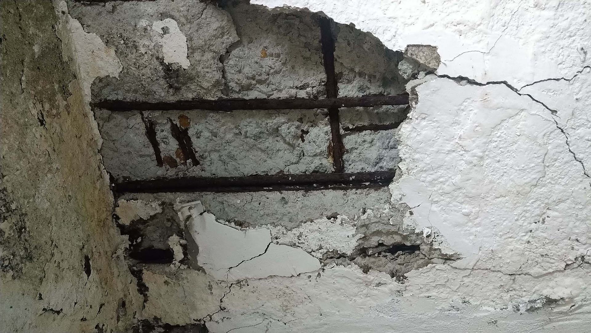 Damaged plaster ceiling with exposed metal bars and cracks in a white, weathered wall