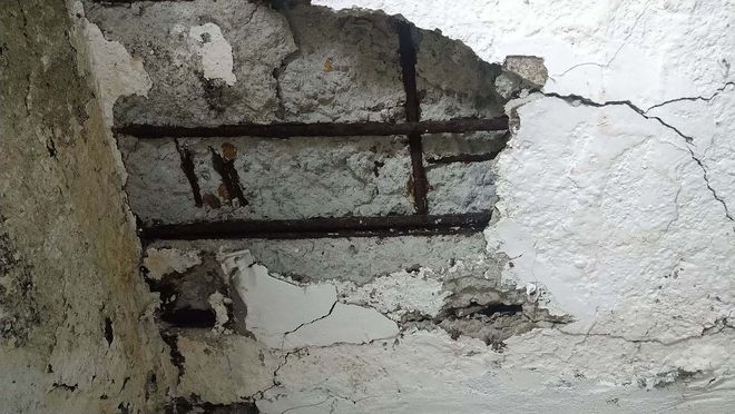 Damaged plaster ceiling with exposed metal bars and cracks in a white, weathered wall