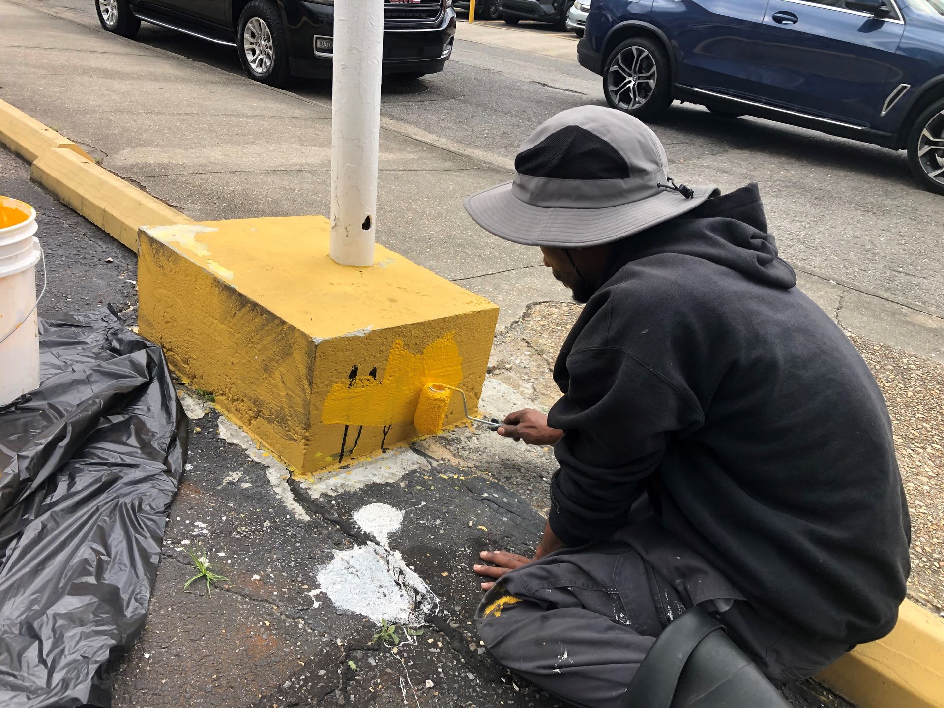 Person kneels beside a yellow utility box, cleaning spilled white powder on a sidewalk near parked cars.