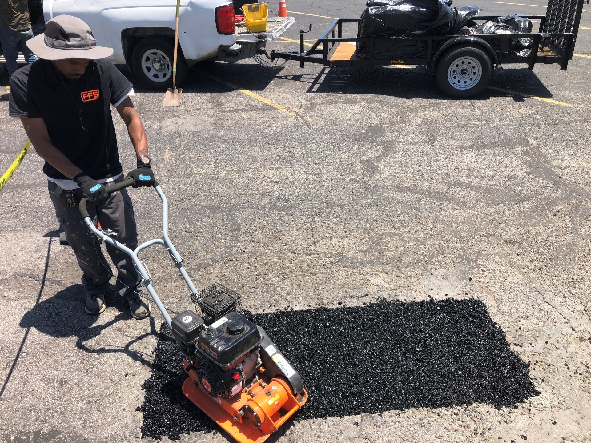 Person using a vibrating plate compactor to pack gravel outdoors, with vehicles in the background