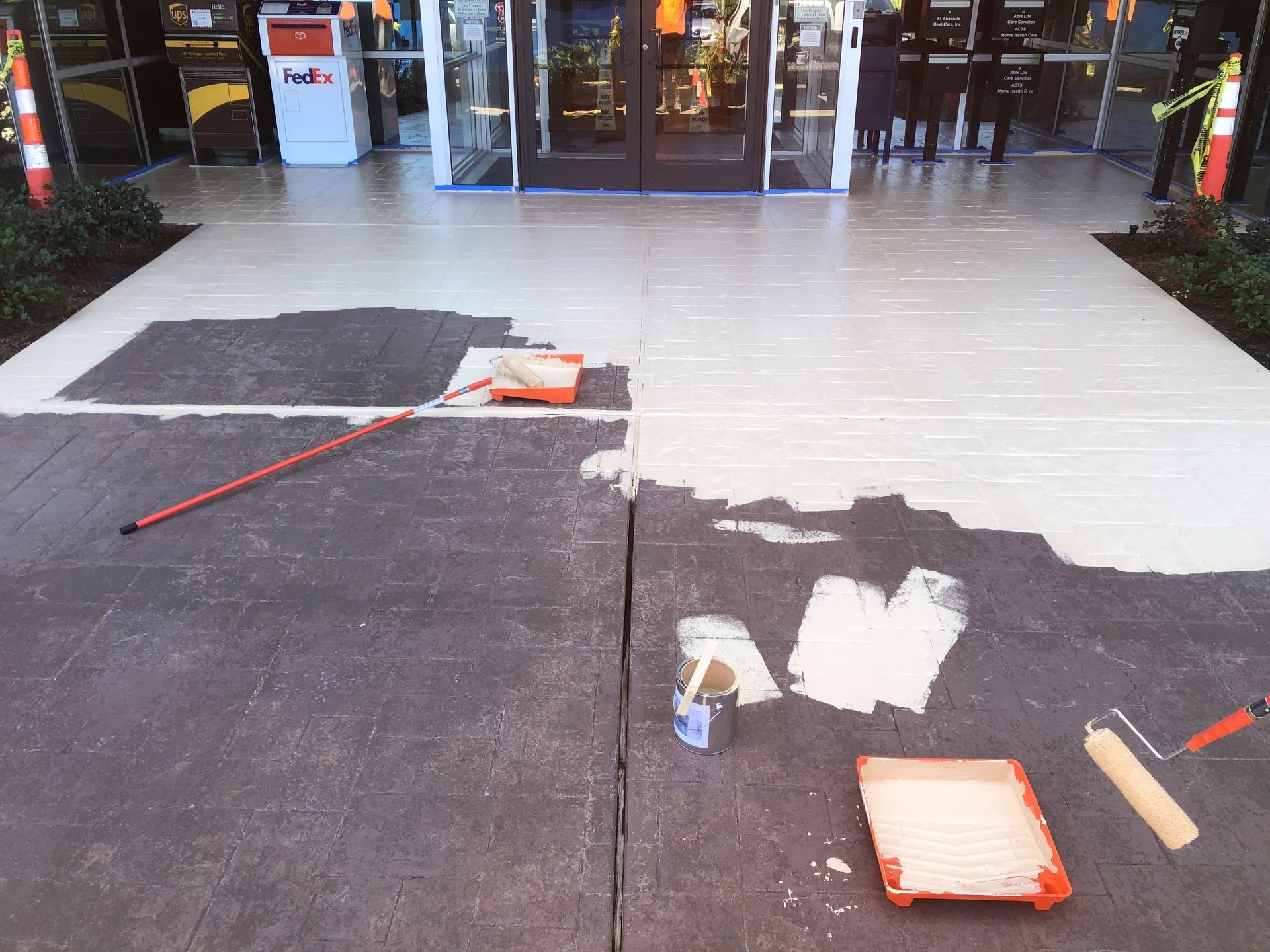 Store entrance with unfinished floor repair, paint rollers, and tools on dark and light flooring.
