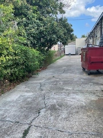 Narrow alley with cracked pavement, trees on the left, and red and white dumpsters near the end.
