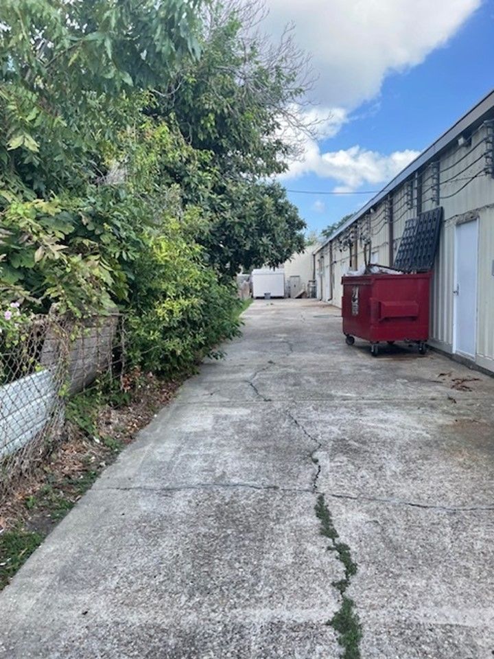 Narrow cracked alley beside industrial buildings, with a red dumpster and trees under a partly cloudy sky.