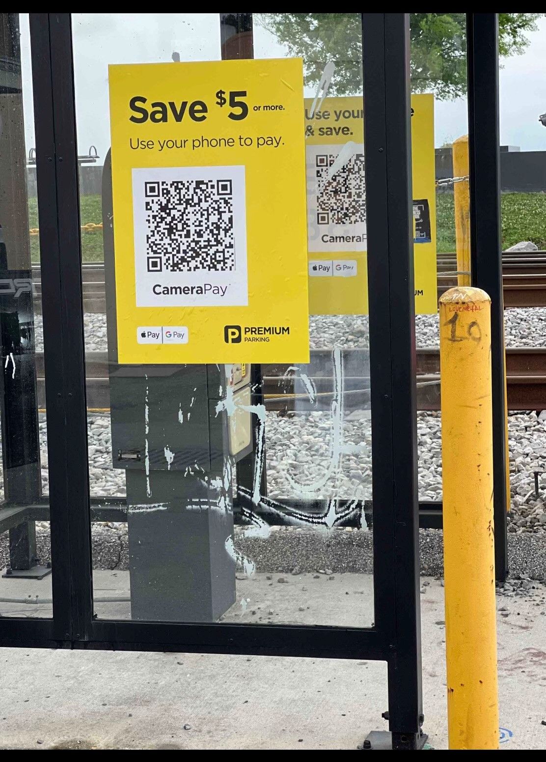Yellow fare-saver sign with QR code on a bus stop shelter beside tracks and a yellow pole.