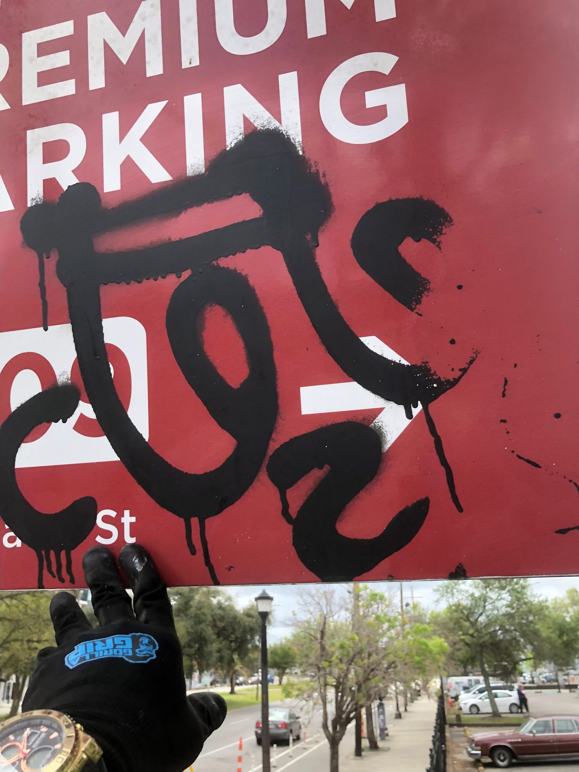 Gloved hand spray-painting black graffiti on a red “Premium Parking” sign near a city street