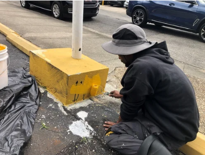 Person crouches beside a yellow curb block, scraping or cleaning white debris on a parking lot pavement.
