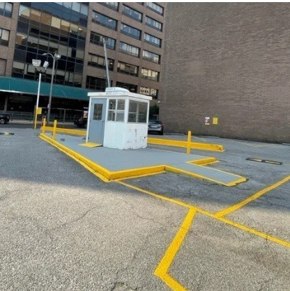Small white utility booth on a marked parking lot, surrounded by yellow painted lines and curbs.