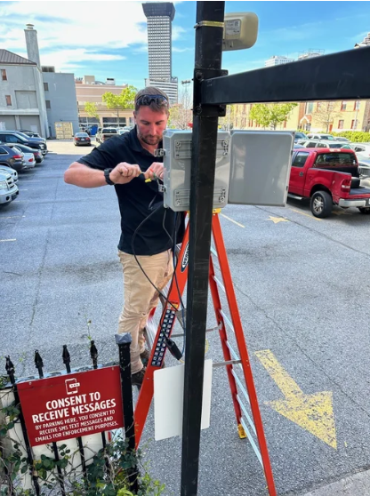 Man adjusting equipment on a metal pole beside a red ladder in a parking lot