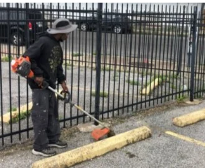 Worker using a string trimmer by a black metal fence in a parking lot.
