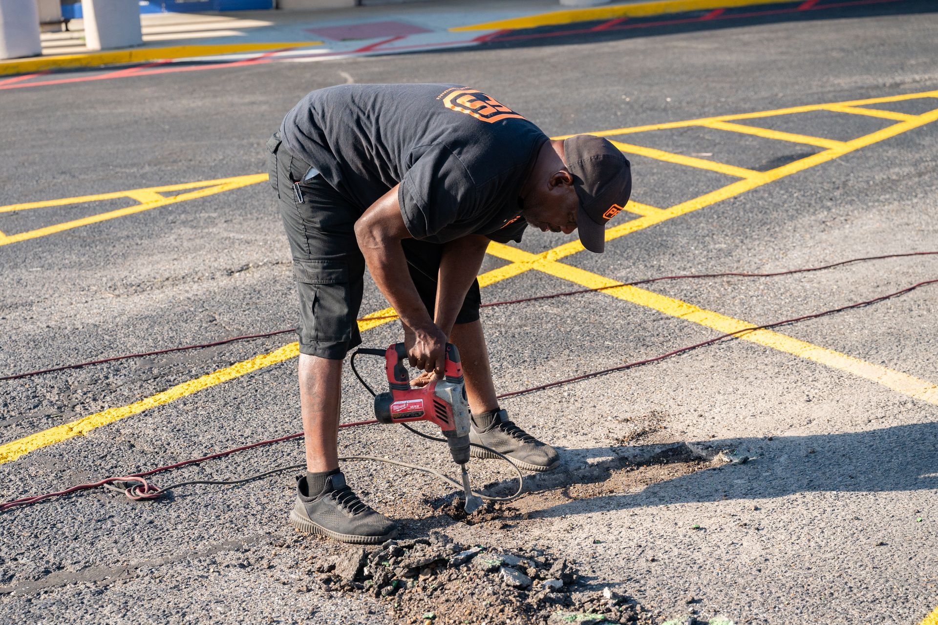 Worker using a red jackhammer to break pavement in a marked parking lot.