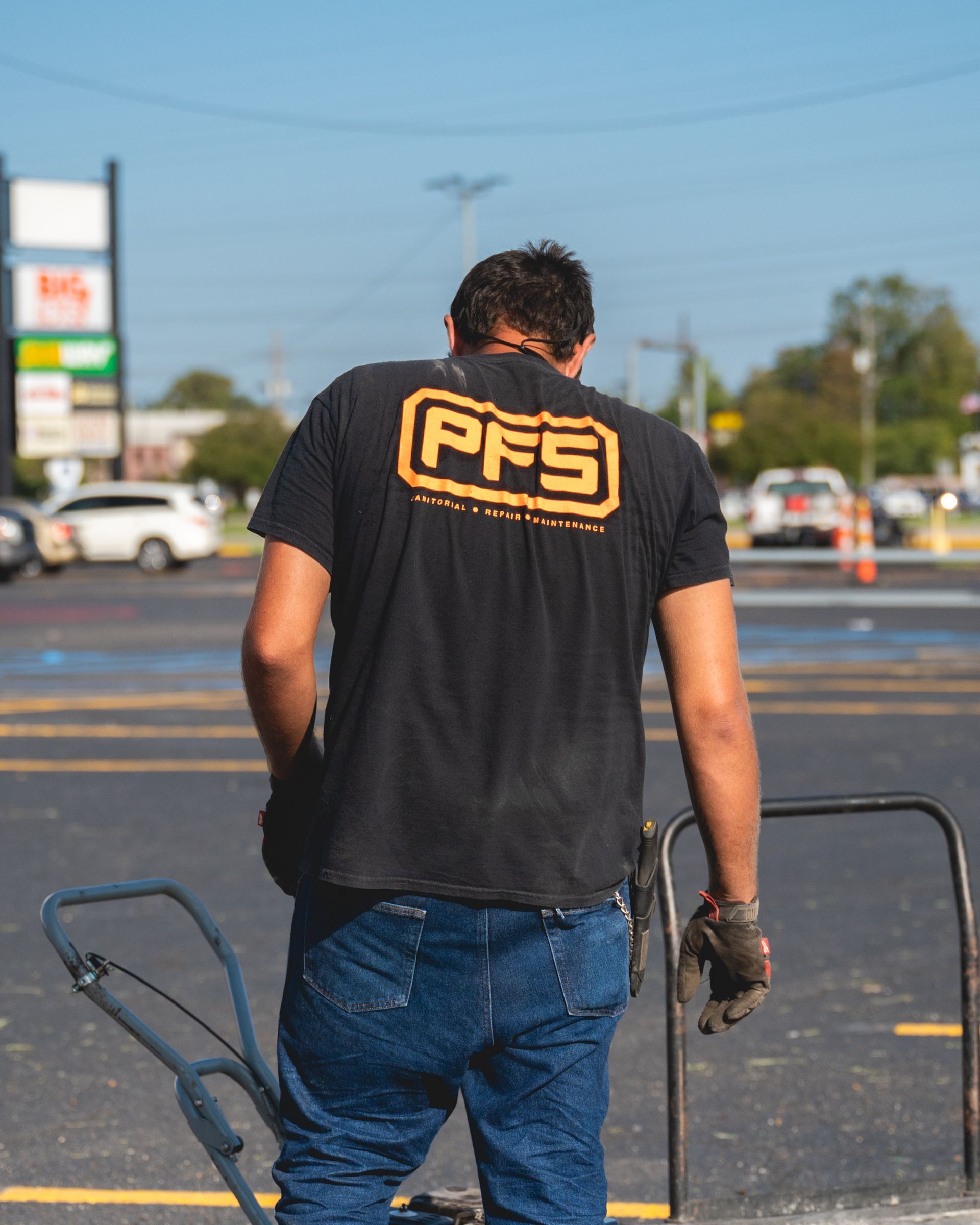 Person in a black PFS shirt walks across a parking lot, carrying gloves and a tool.