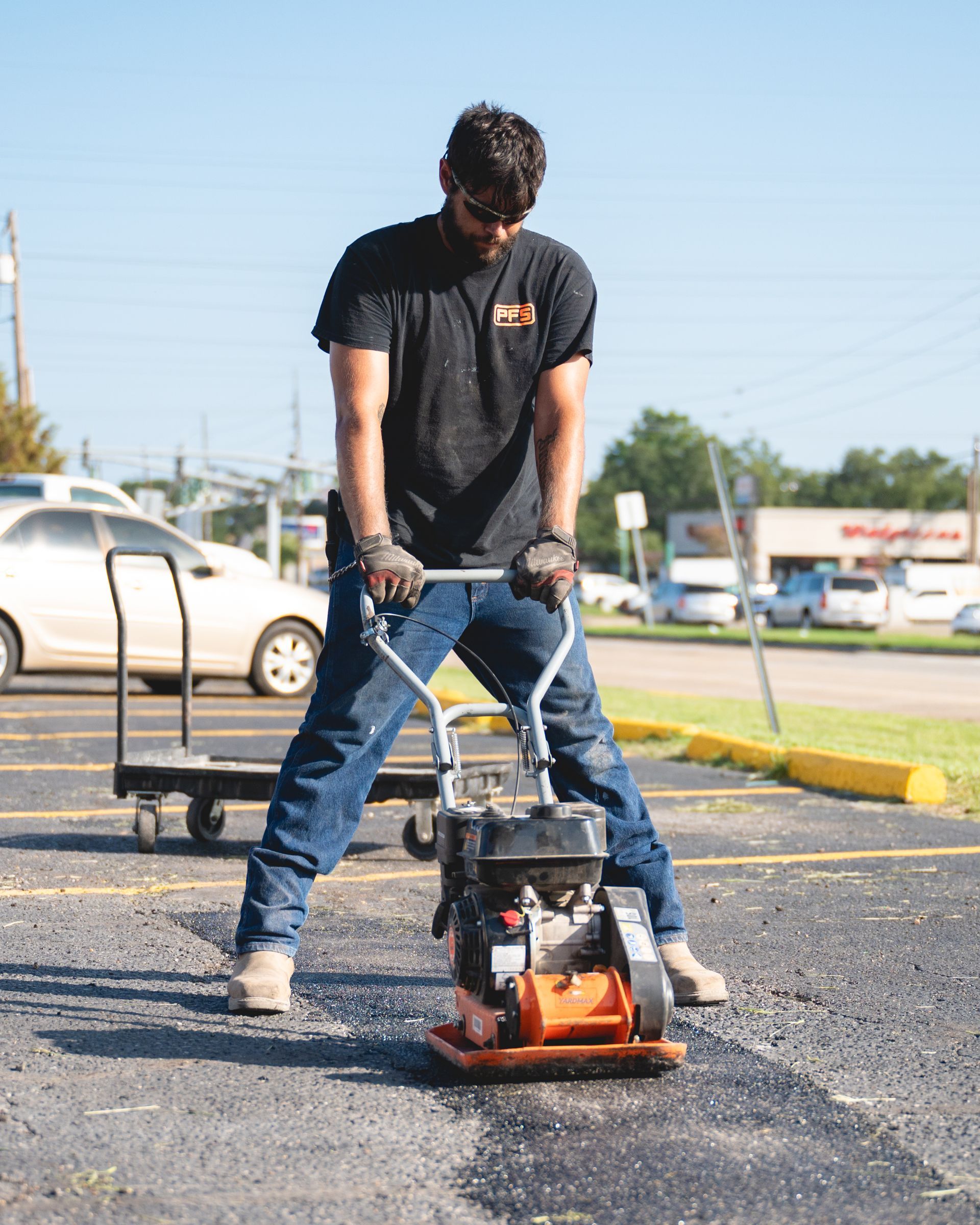Person operating a plate compactor on gravel in a parking lot, with a car and trailer in the background