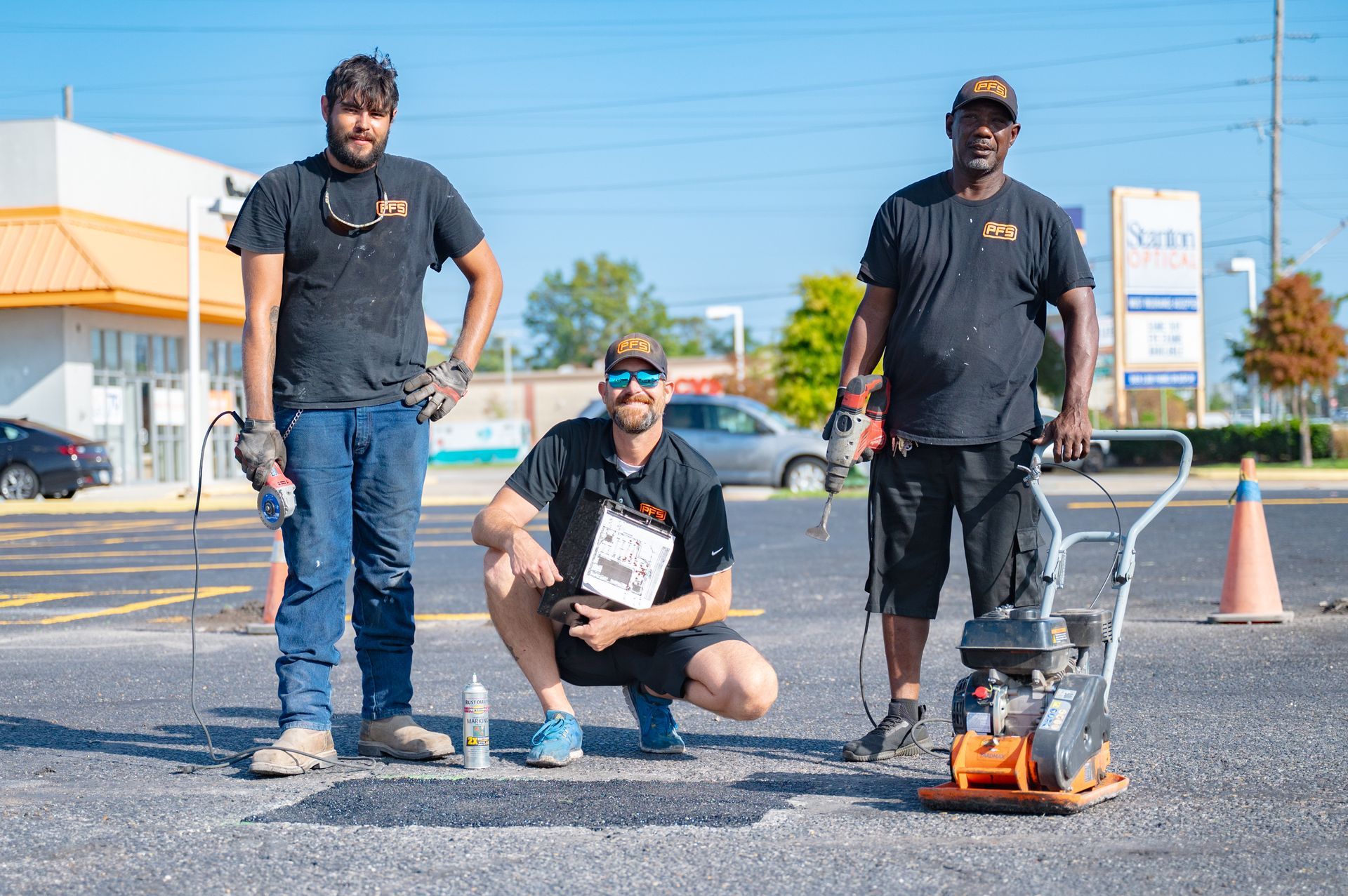 Three workers posing beside a small road machine in a parking lot under a clear blue sky