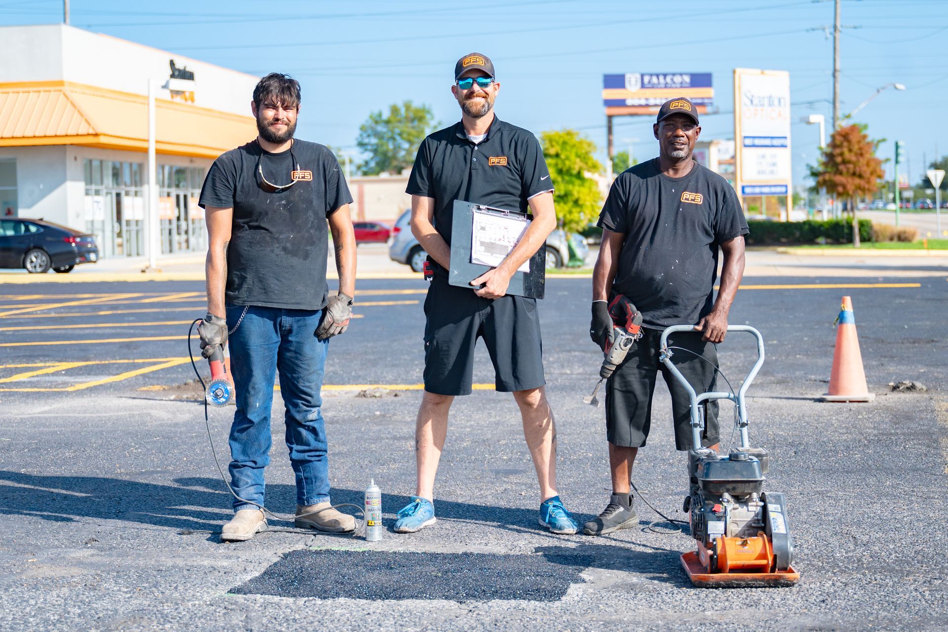 Three workers in black shirts stand with surveying equipment on a sunlit roadside.