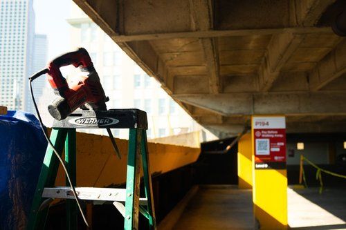Tilted red chair on a metal stand under a concrete overpass, with a yellow pillar and walkway nearby.