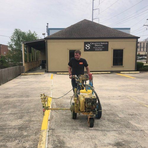 Worker pushing a yellow concrete grinder on a parking lot beside a beige building