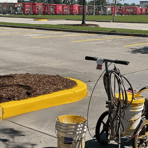 Parking lot with a line-striper cart and yellow paint buckets beside a freshly painted curb.