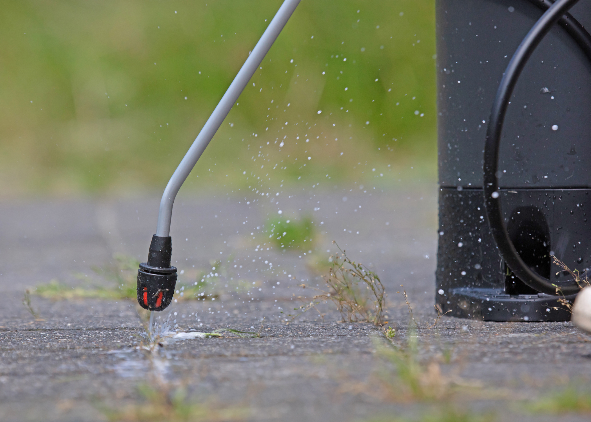 A hose nozzle spraying water onto pavement near a black container, with a blurred green background.