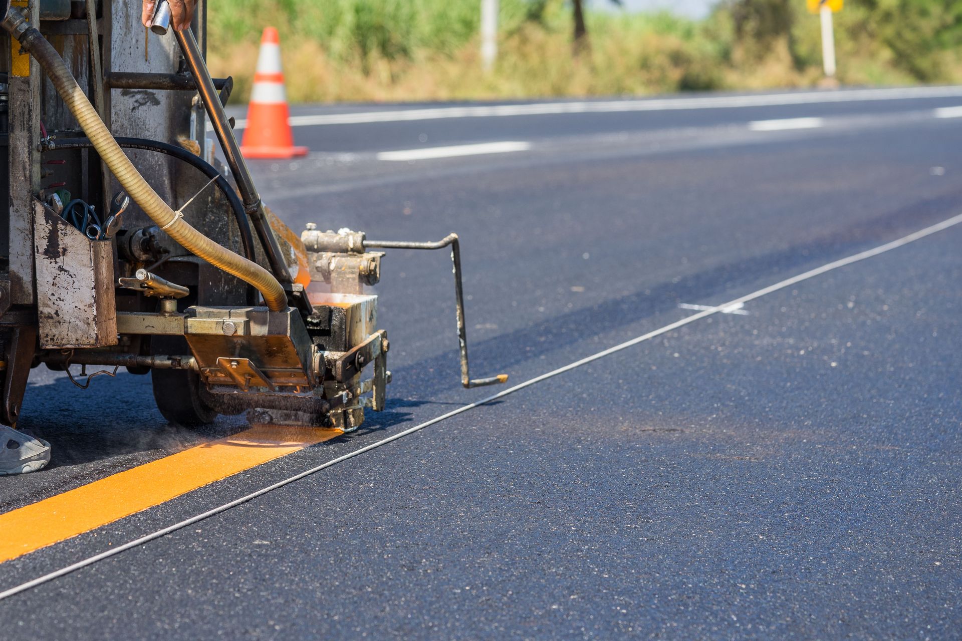 Roadwork machine painting a yellow line on asphalt highway near an orange traffic cone.