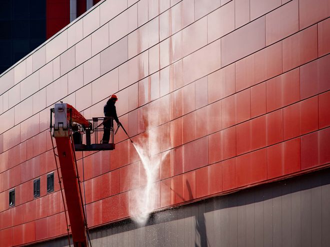Worker pressure-washing a red building facade from a lift platform