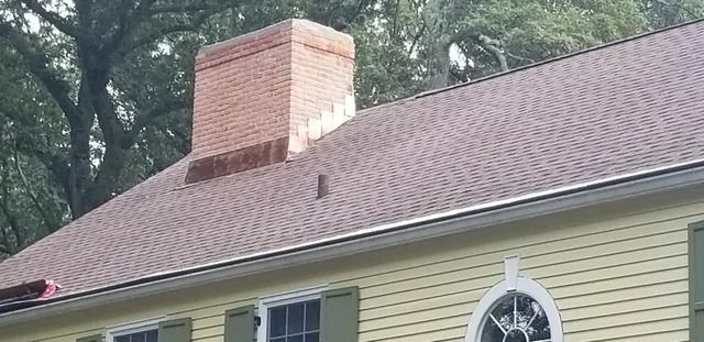 Brick chimney on a yellow house with a shingled roof and decorative window trim