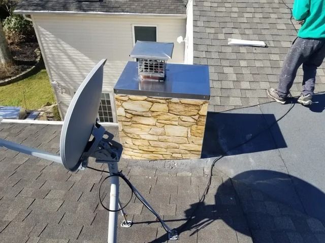 Roof with satellite dish, solar panel, and a person standing on gray shingles near a stone chimney.