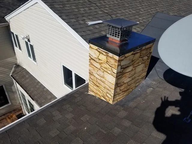 Rooftop with a stone chimney, dark shingle roof, and neighboring house siding under daylight