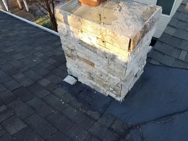 Stone chimney on a dark shingled roof with visible damage and cracked mortar
