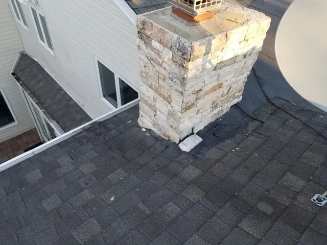 Rooftop shingle repair around a brick chimney beside a light gray house exterior