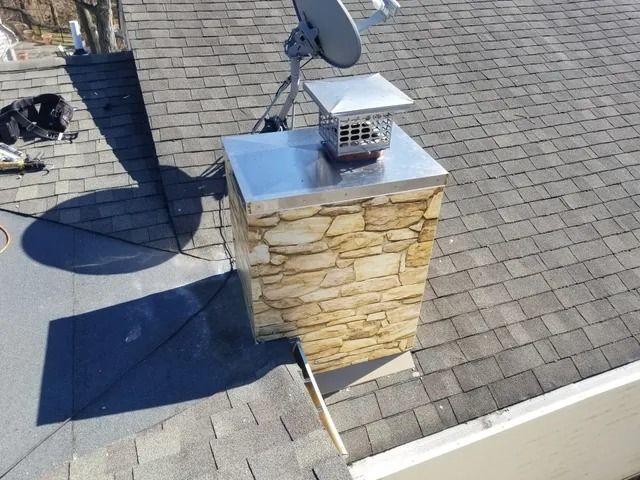 Rooftop stone chimney with metal cap and satellite dish on gray shingle roof in sunlight