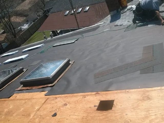 Roof under construction with skylights, gray membrane, and a worker kneeling near the top right