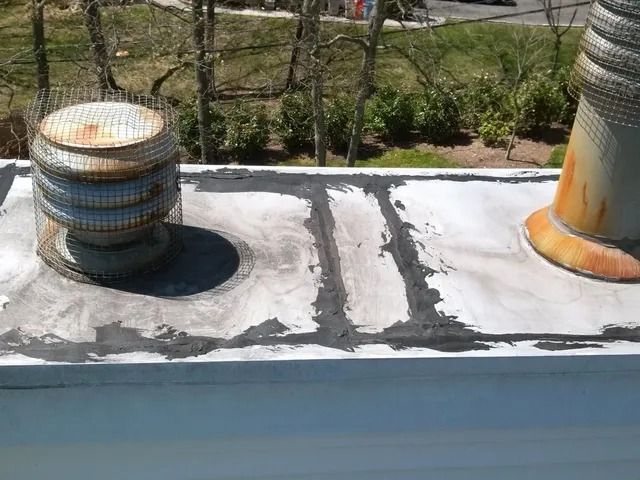 Two rooftop vents on a weathered flat roof, one blue-gray, one orange-rusted, with a fence and greenery behind