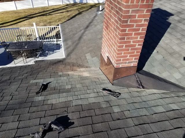 Rooftop with dark shingles, brick chimney, and patio furniture near a white railing.