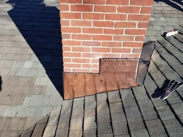 Roof shingles around a brick chimney with brown flashing at the base