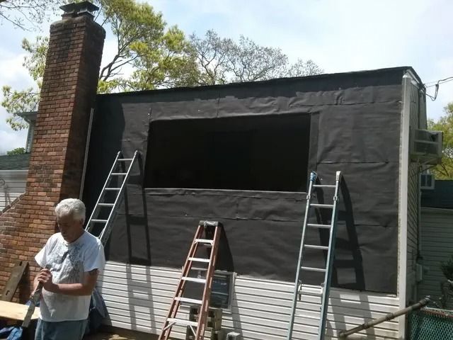 House exterior with black tarp covering an upper wall; ladders lean against it beside a brick chimney.