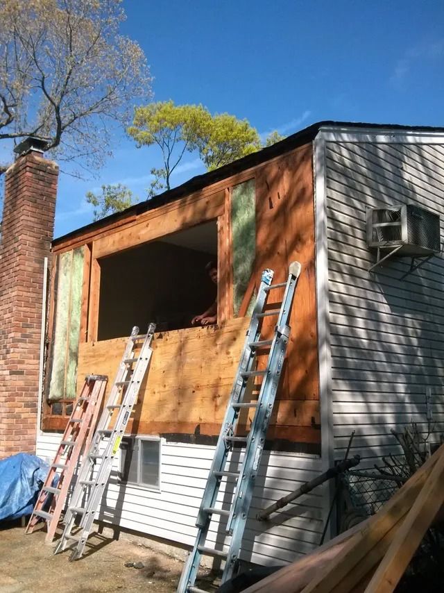 House exterior under renovation with orange plywood, ladders, and gray siding under a blue sky
