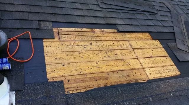 Roofing shingles around a plywood patch on a dark shingled roof, with an orange cord on the left.
