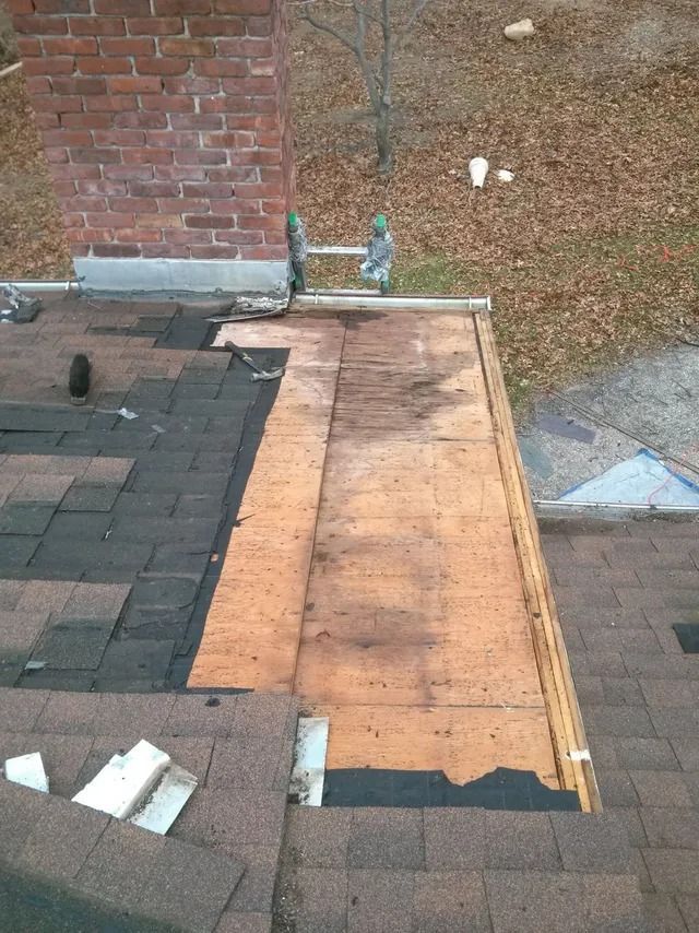 Roof repair with exposed plywood beside dark shingles and a brick chimney