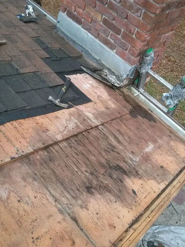 Damaged roof with plywood, shingles, and exposed flashing beside a brick wall