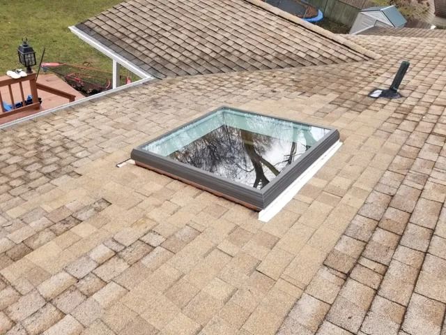 Rooftop with a square skylight on shingle shingles, with trees reflected in the glass.