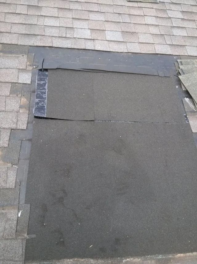 Flat black roofing patch on a shingled roof, with gray shingles around the edges.