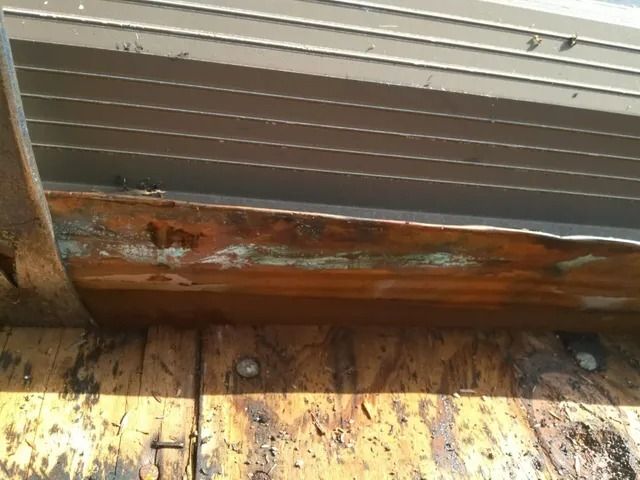 Rusty metal beam under a corrugated roof, with weathered wooden floorboards below.