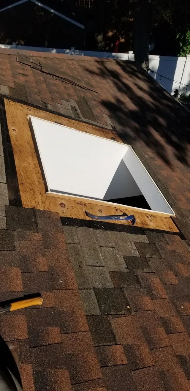 Rooftop with a square open skylight framed in wood and surrounded by brown shingles.