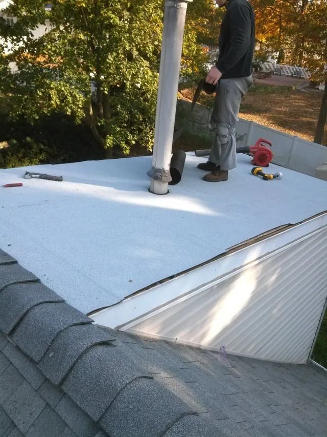 Worker on a white roof installing roofing around a vent pipe, with tools nearby.