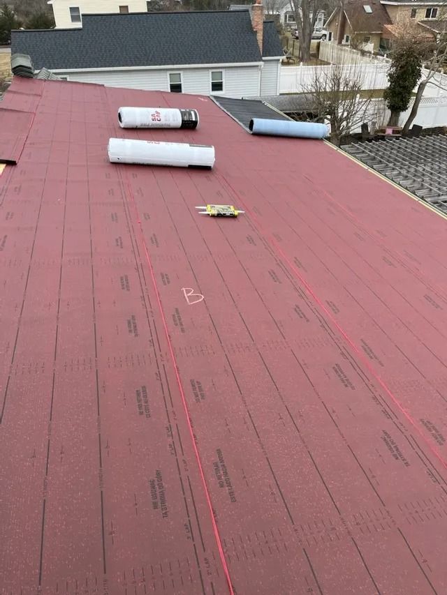 Red roofing underlayment on a sloped roof with rolled materials near the ridge and houses in the background