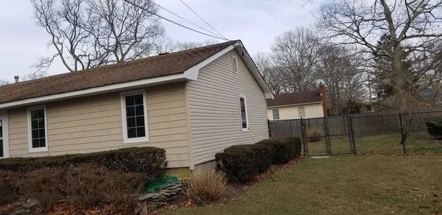 Single-story beige house with brown roof and shrubs beside a fenced yard on a cloudy day